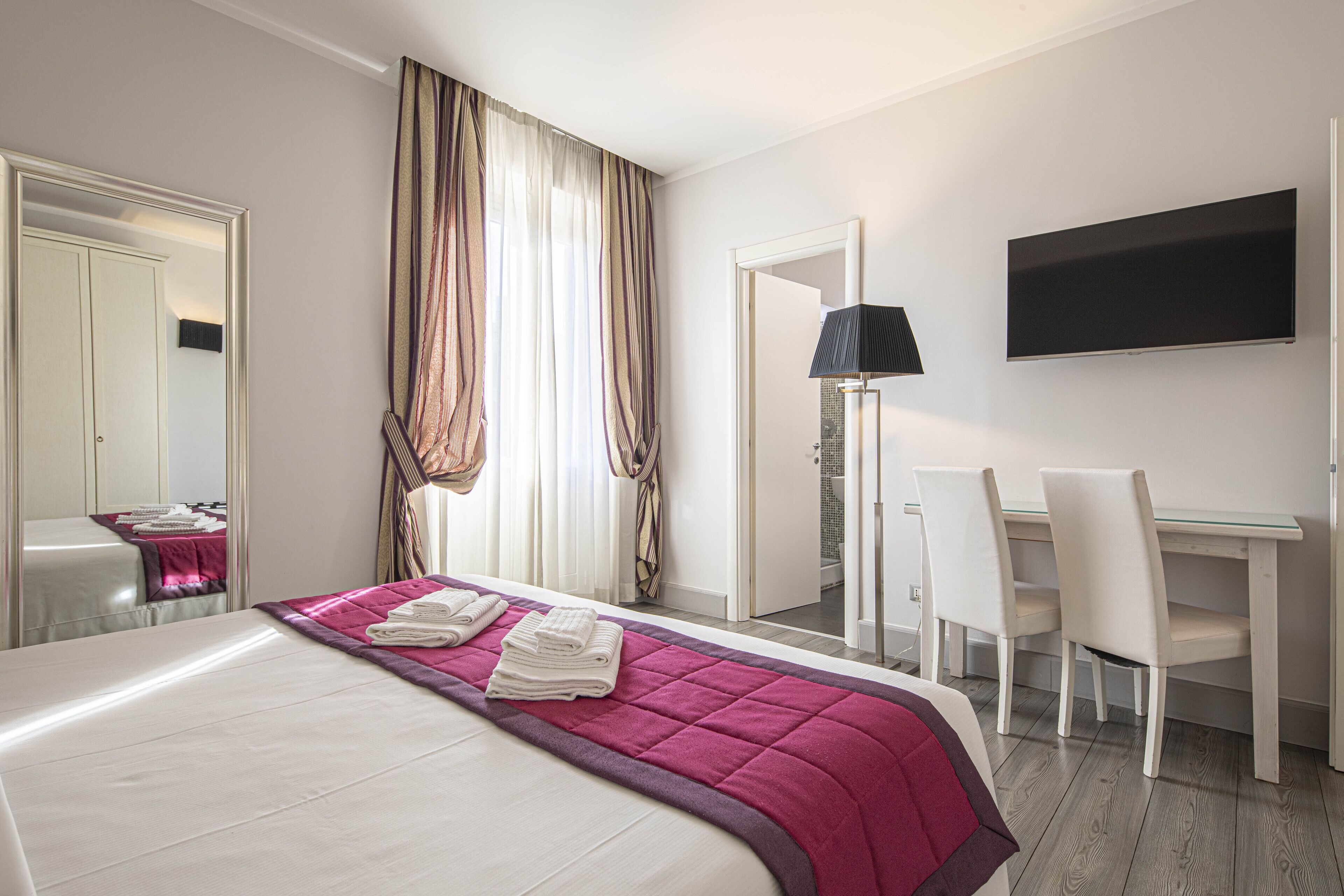 Une chambre d’hôtel avec un lit, un bureau, deux chaises, une télévision et une porte de salle de bain.