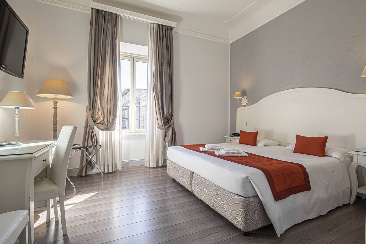 Habitación de hotel con cama, escritorio, silla, televisor y ventana con cortinas.