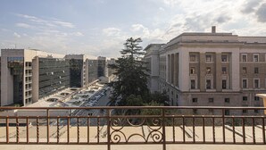 A view from a balcony with a metal railing, overlooking a city street with buildings, a tree, and a clear sky. - Suite Castrense (Rome)