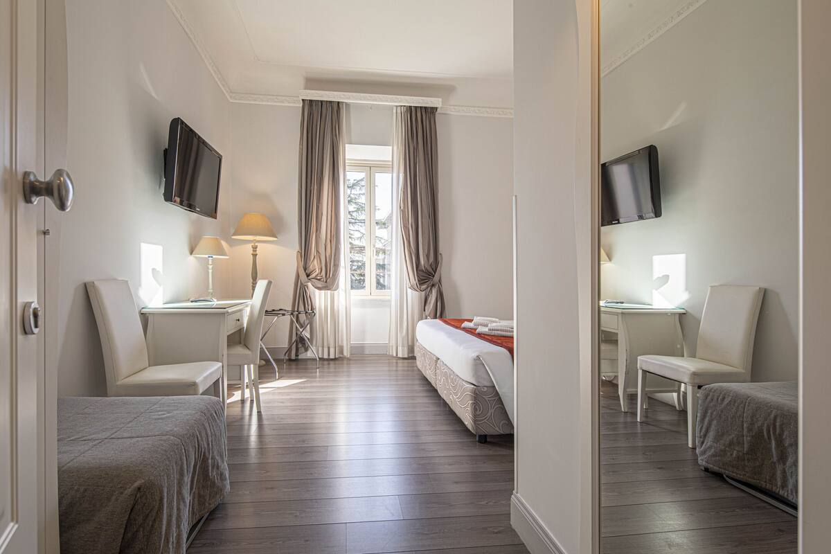 Una habitación de hotel con cama, dos sillas, escritorio, televisor y ventana con cortinas.