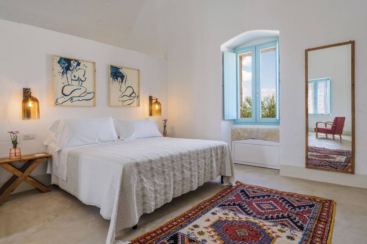 A bedroom with a bed, two wall-mounted artworks, a bedside table with a lamp, a window with a view, and a large mirror.