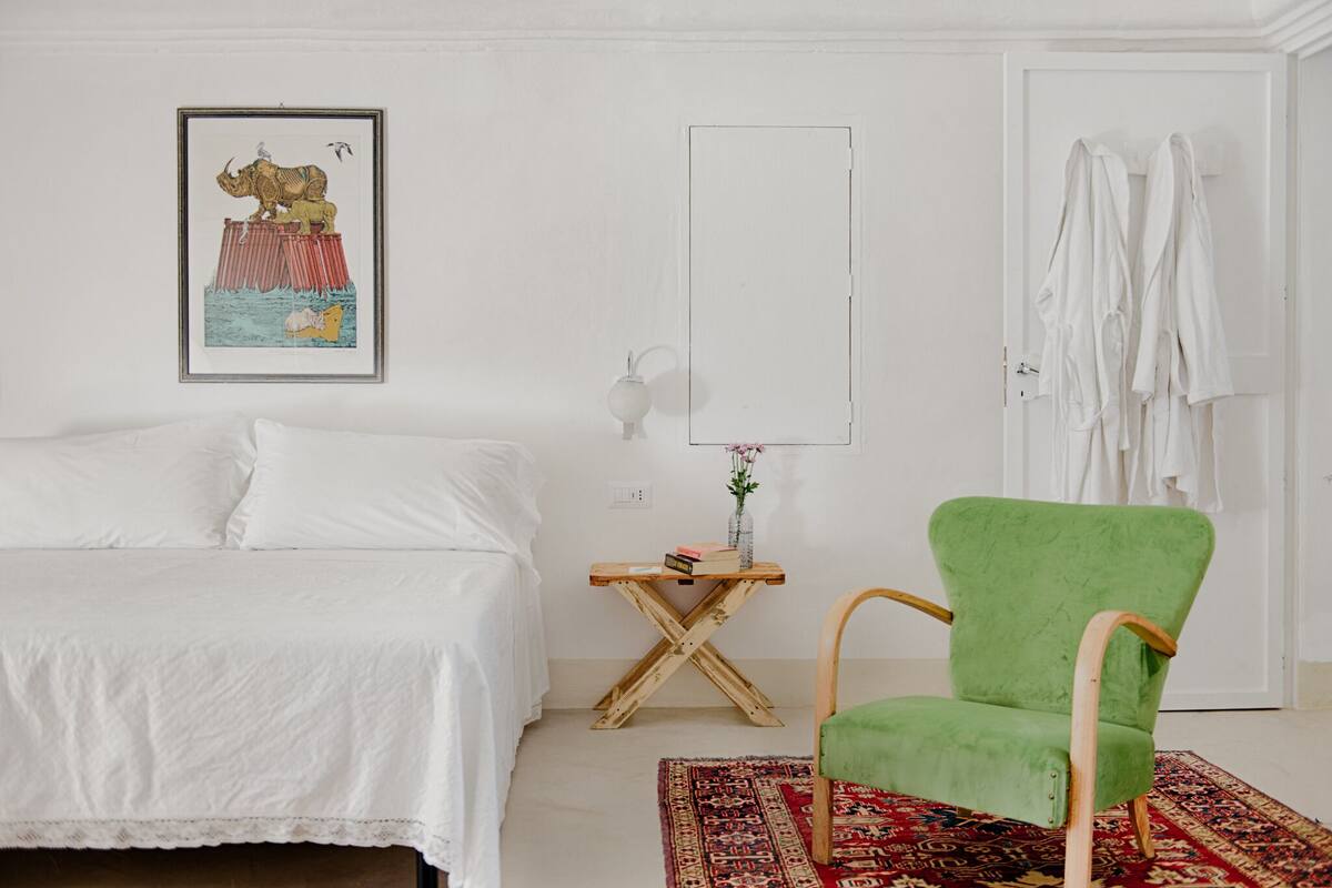 A bedroom with a bed, a green chair, a small table with a vase, and a framed picture on the wall.