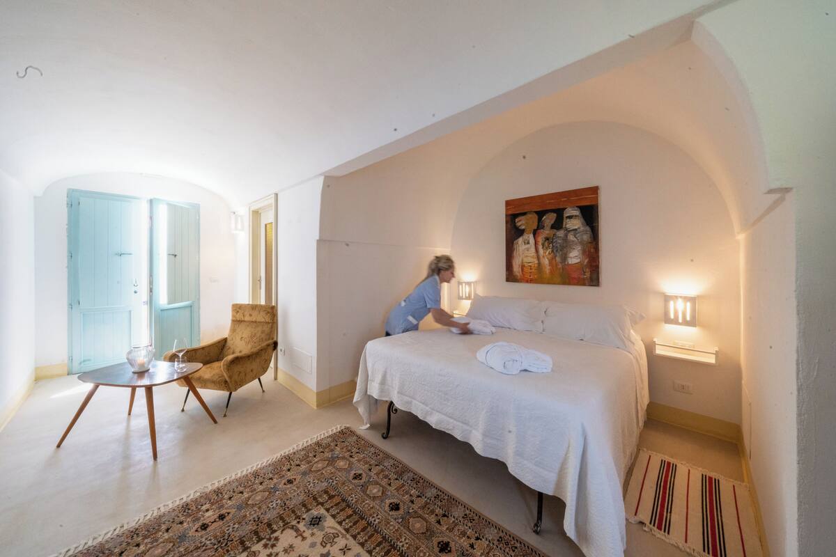 A bedroom with a bed, a chair, a table, a painting, and a lamp.