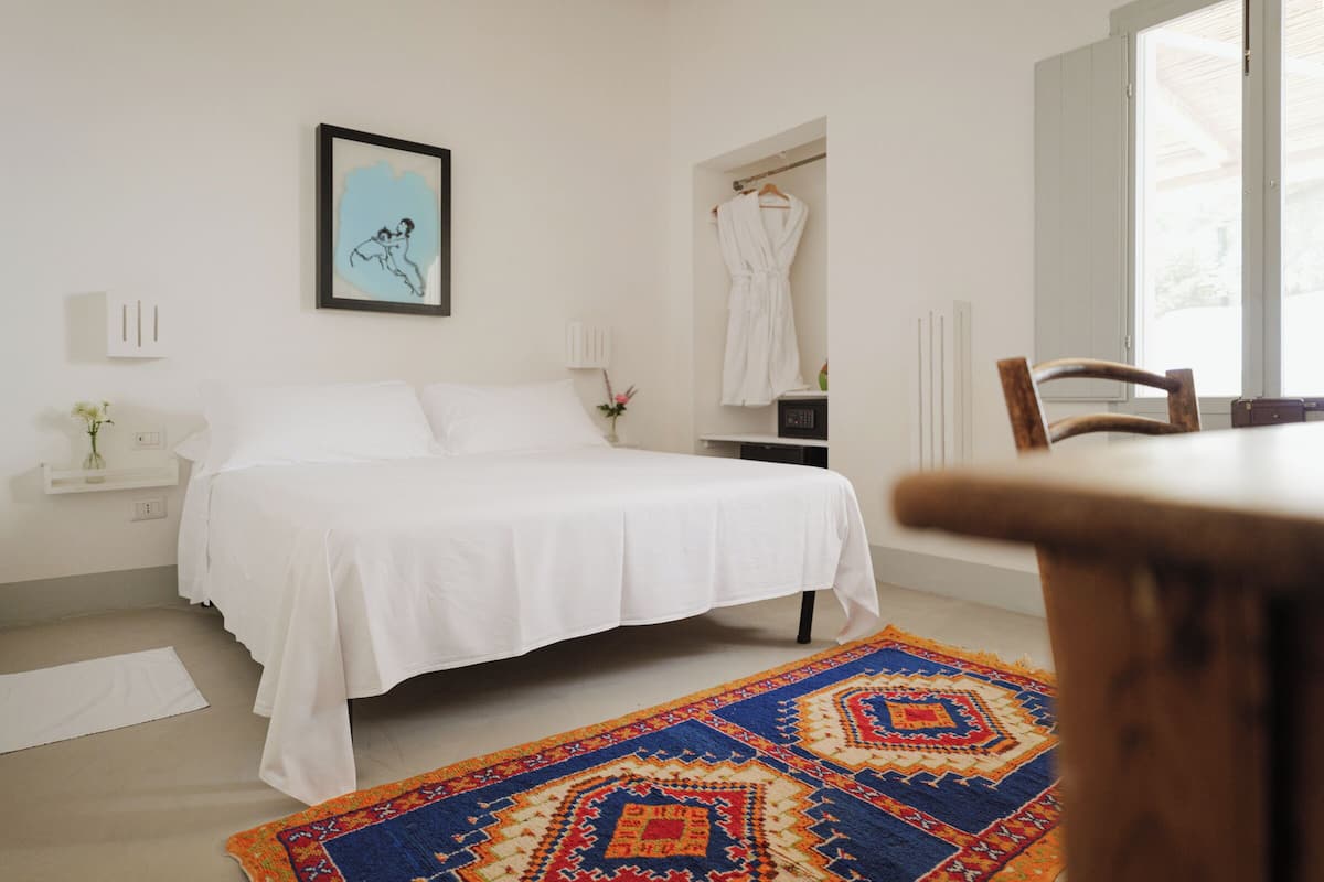 A bedroom with a bed, a bedside table, a chair, a window, and a patterned rug.