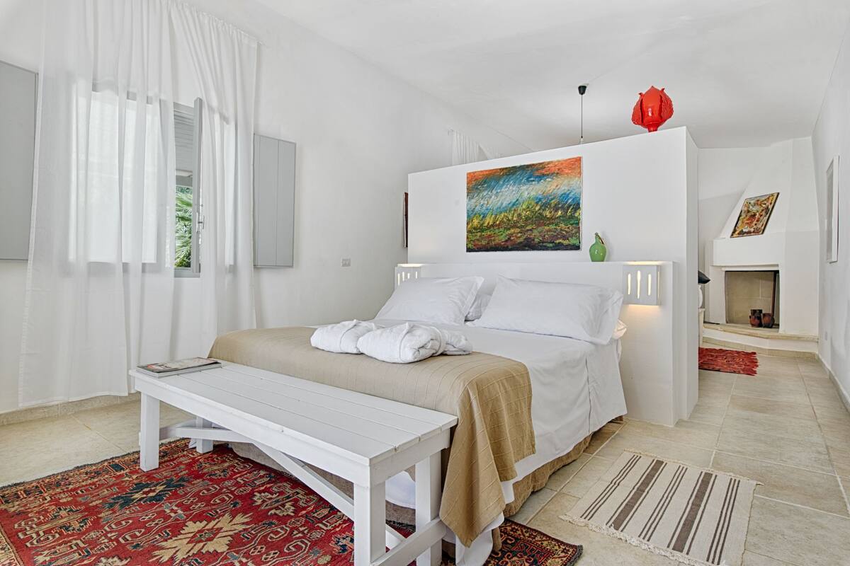 A modern hotel room with a bed, white furniture, a painting on the wall, and a red decorative item.
