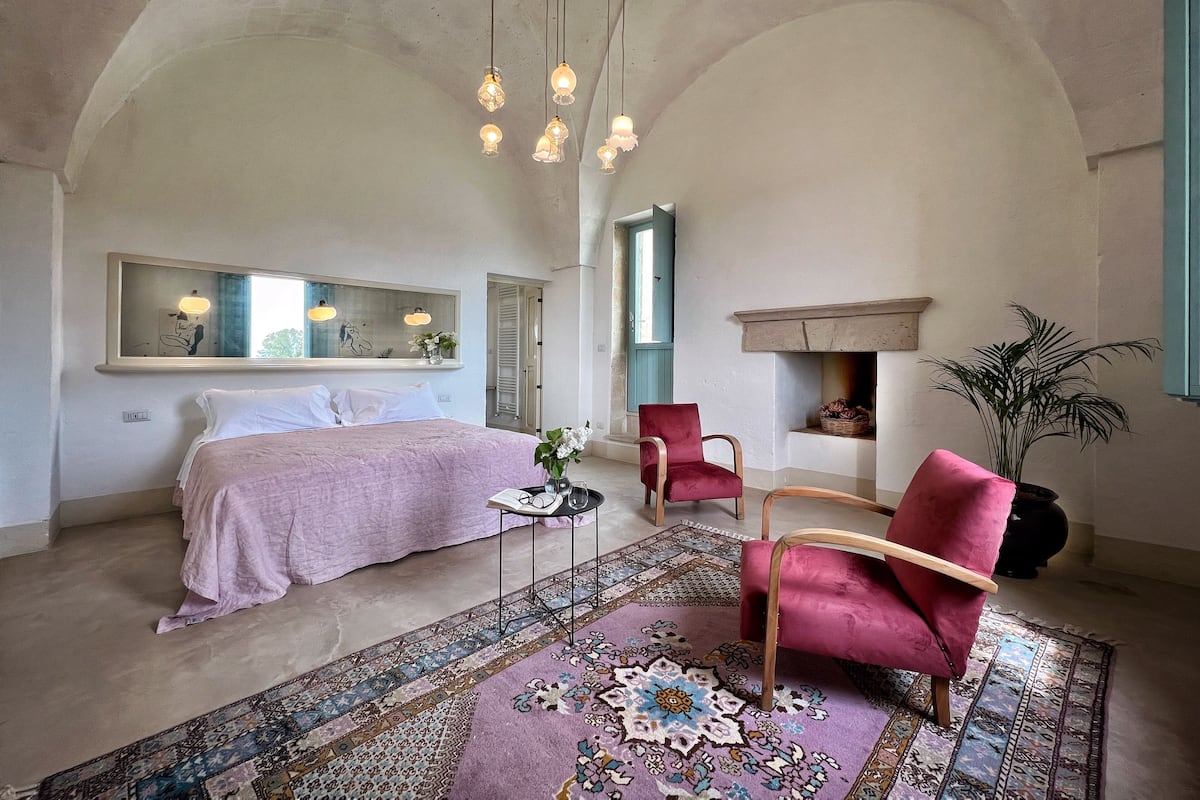 A bedroom with a bed, a chair, a mirror, a fireplace, and a hanging light fixture.