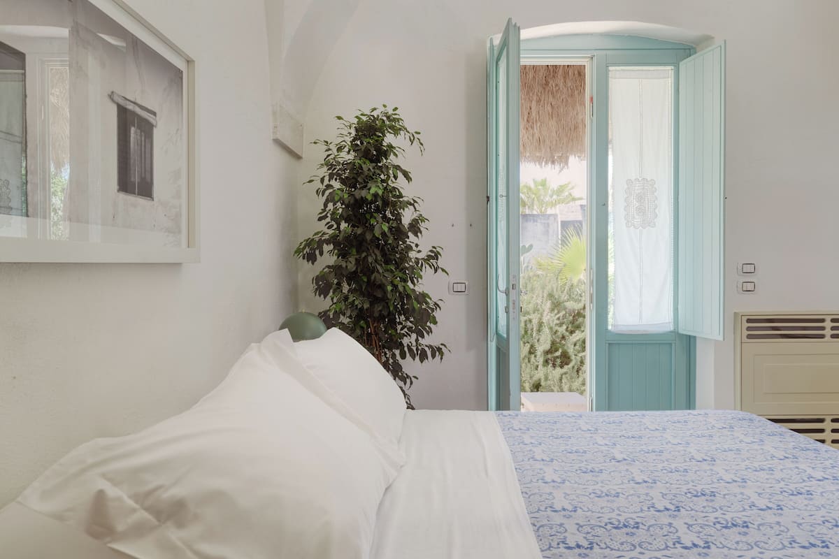 A bedroom with a bed, a potted plant, a window, and an open door leading outside.