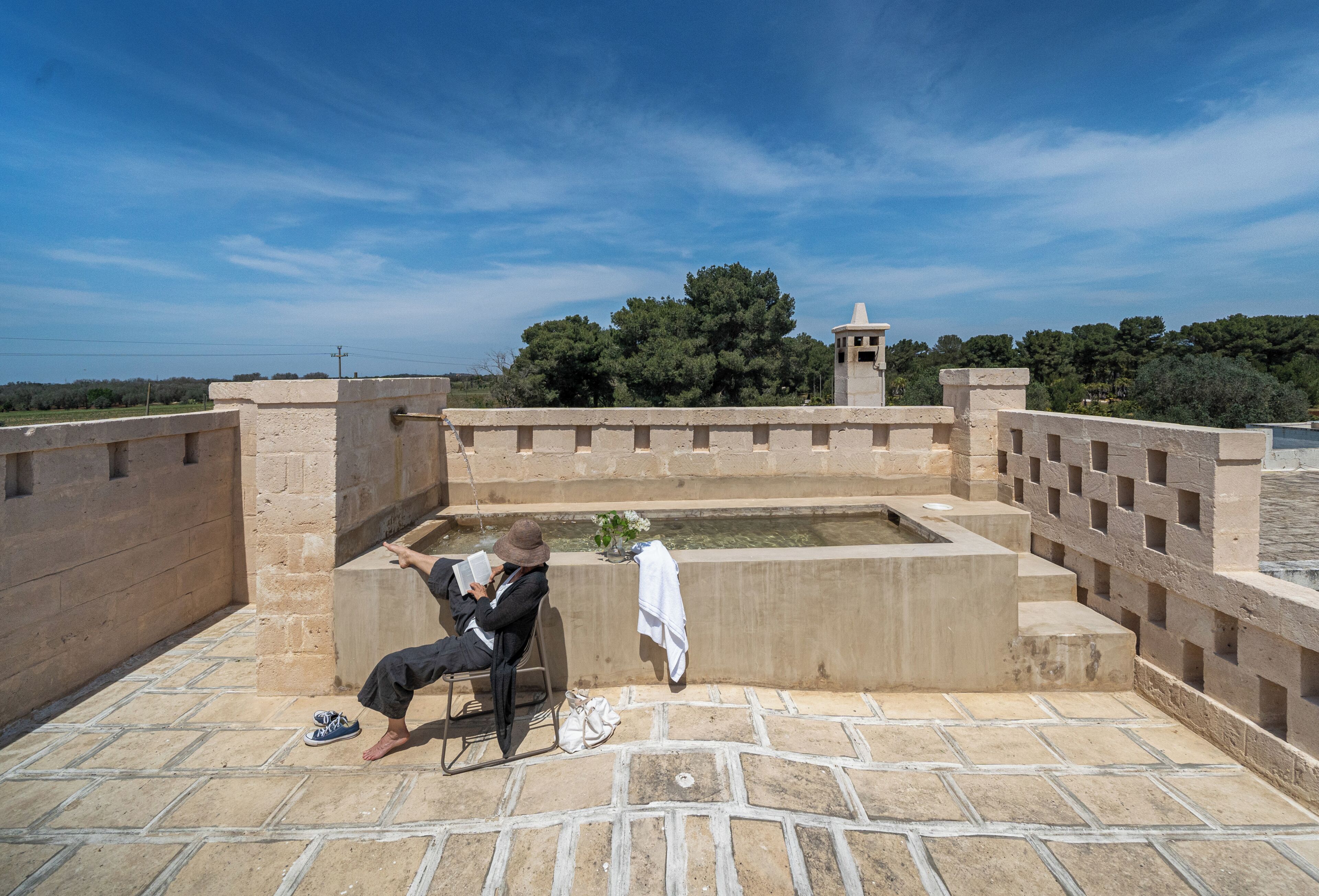 A person sitting on a chair by a stone wall with a small fountain, a clear sky, and trees in the background.