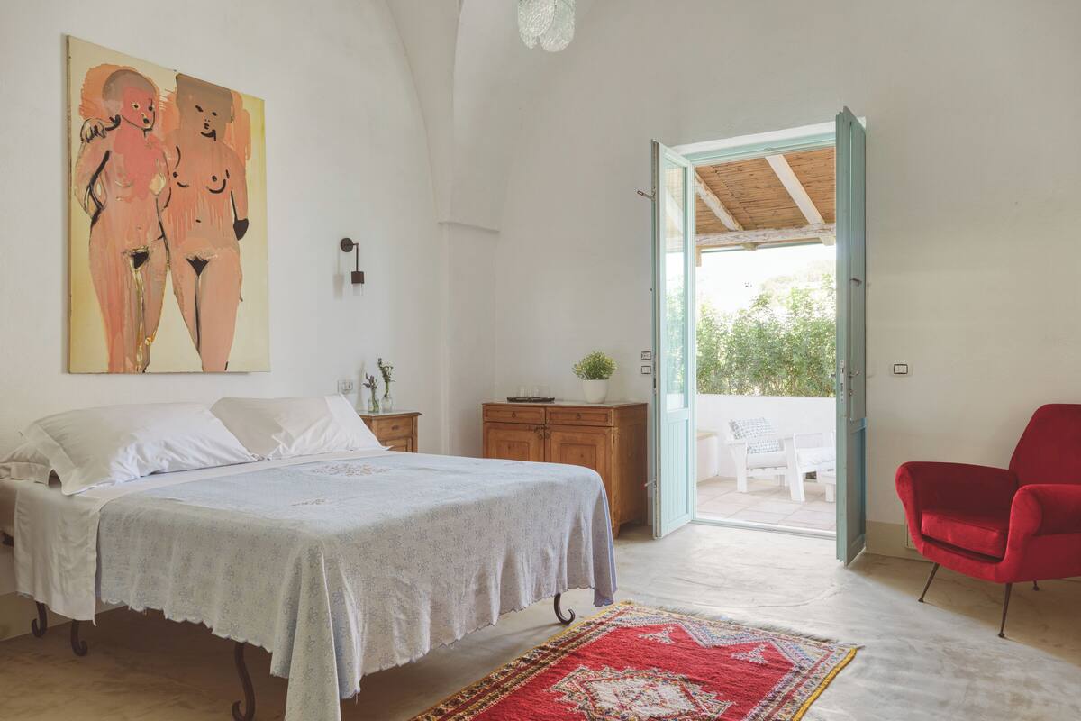 A bedroom with a bed, a red chair, a painting, a wooden cabinet, and a view through an open door.