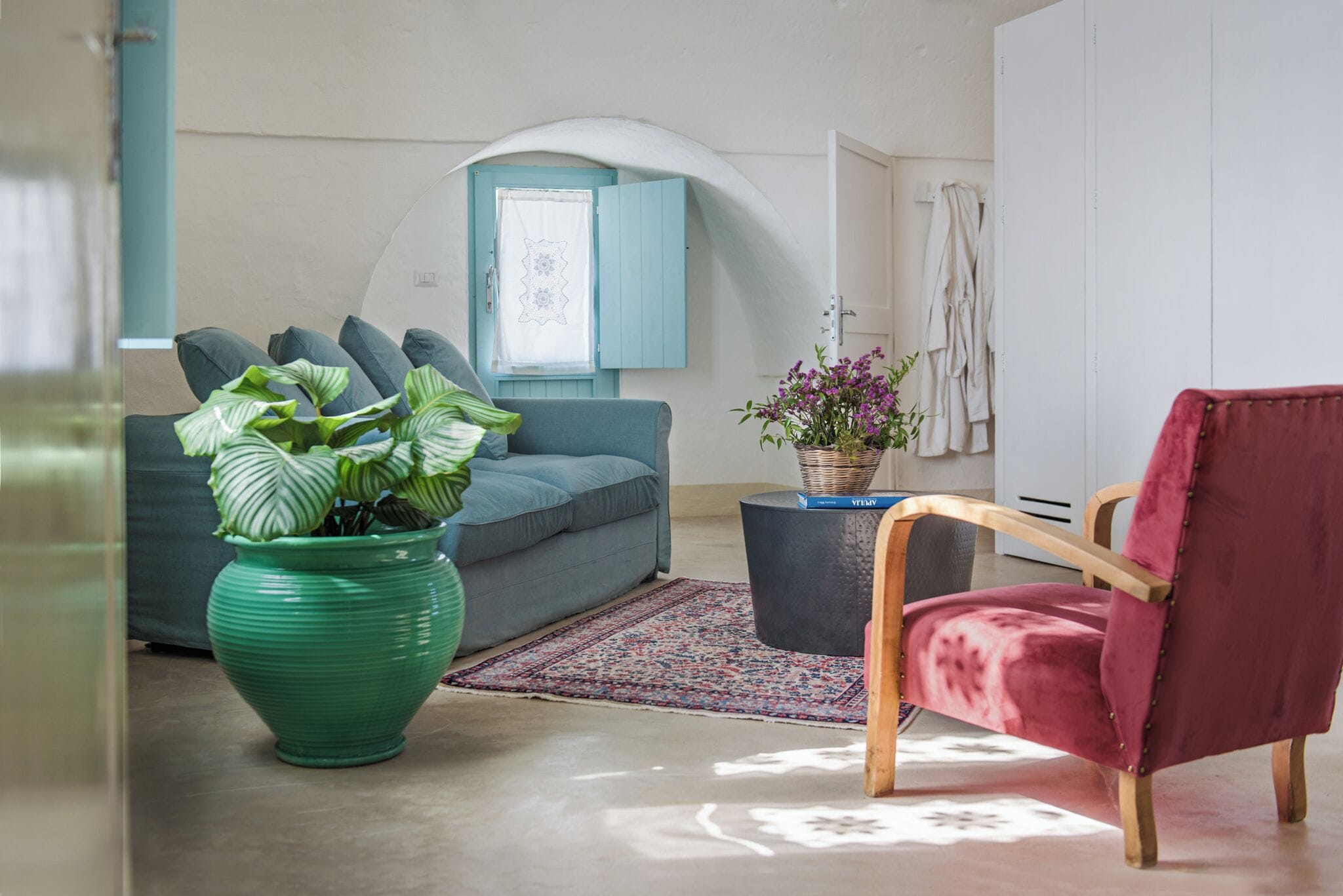 En stue med sofa, rød lænestol, grøn plante og et vindue med blå skodder.