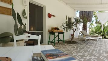Un área de patio techada con una mesa blanca, una silla, una pequeña mesa de madera con cajas, una planta en maceta y una silla de descanso.