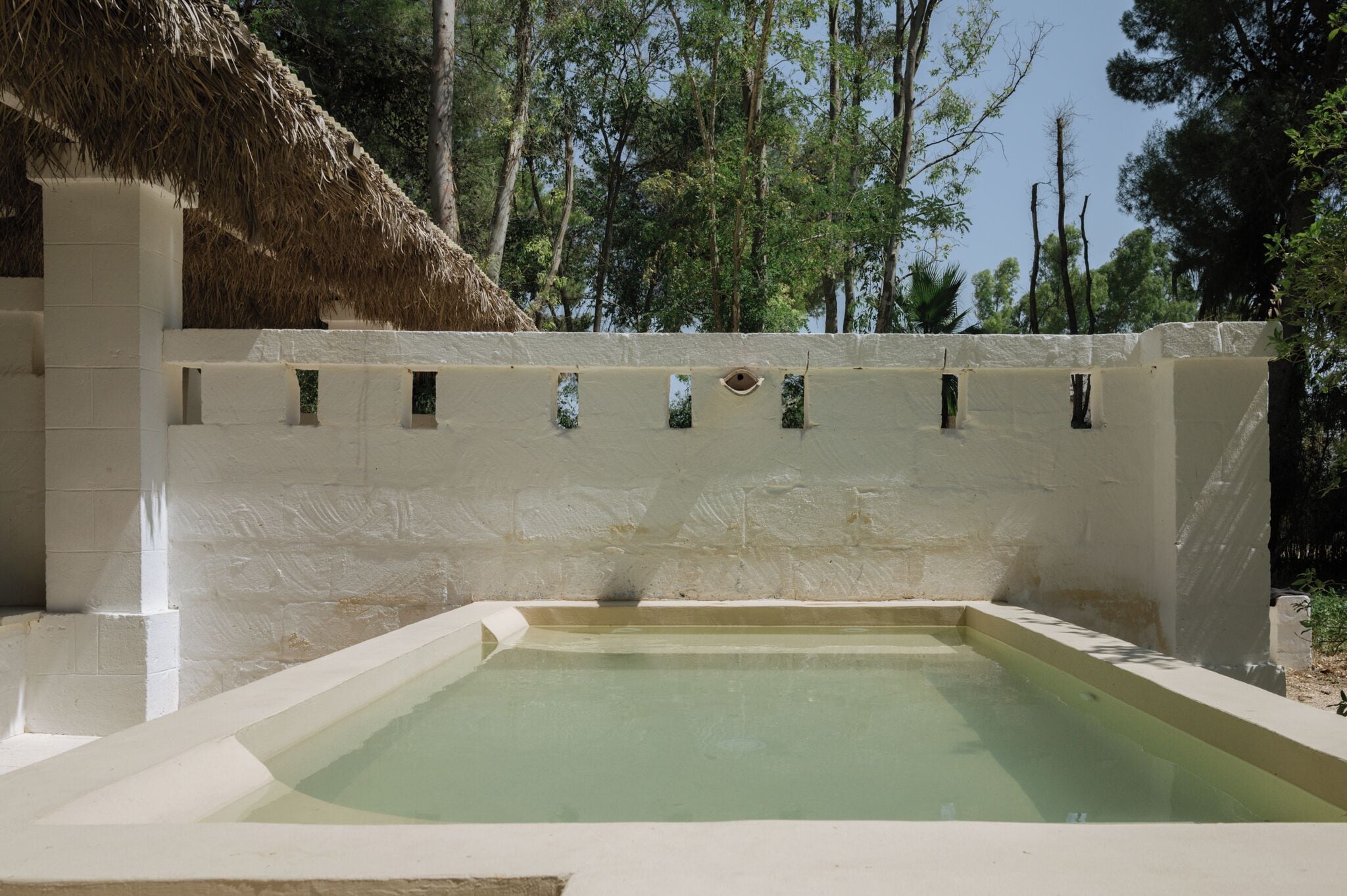 Ein Pool mit überdachter Struktur aus Schilfdächern, umgeben von einer weißen Mauer mit kleinen Fenstern.