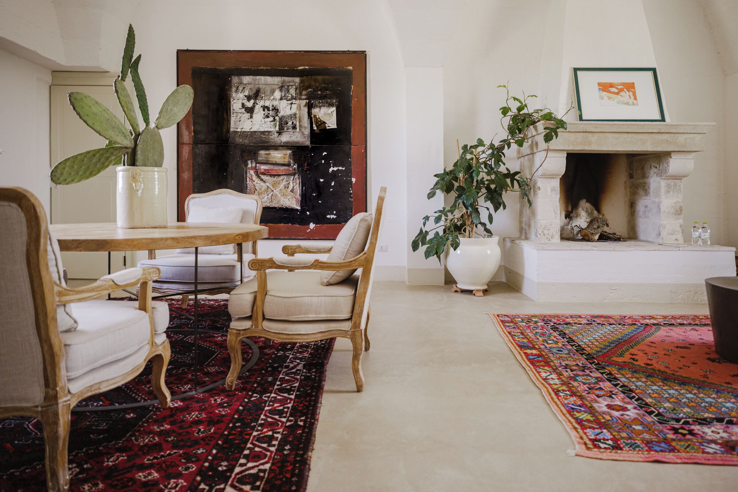 A room with a fireplace, a dining table with chairs, a large plant, and a colorful rug.