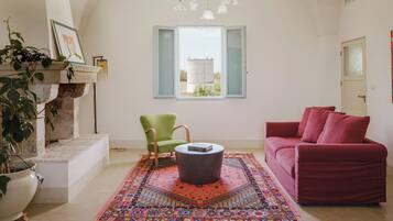 A living room with a red sofa, a green chair, a round coffee table, a colorful rug, a stone fireplace, and a window with an open shutter.
