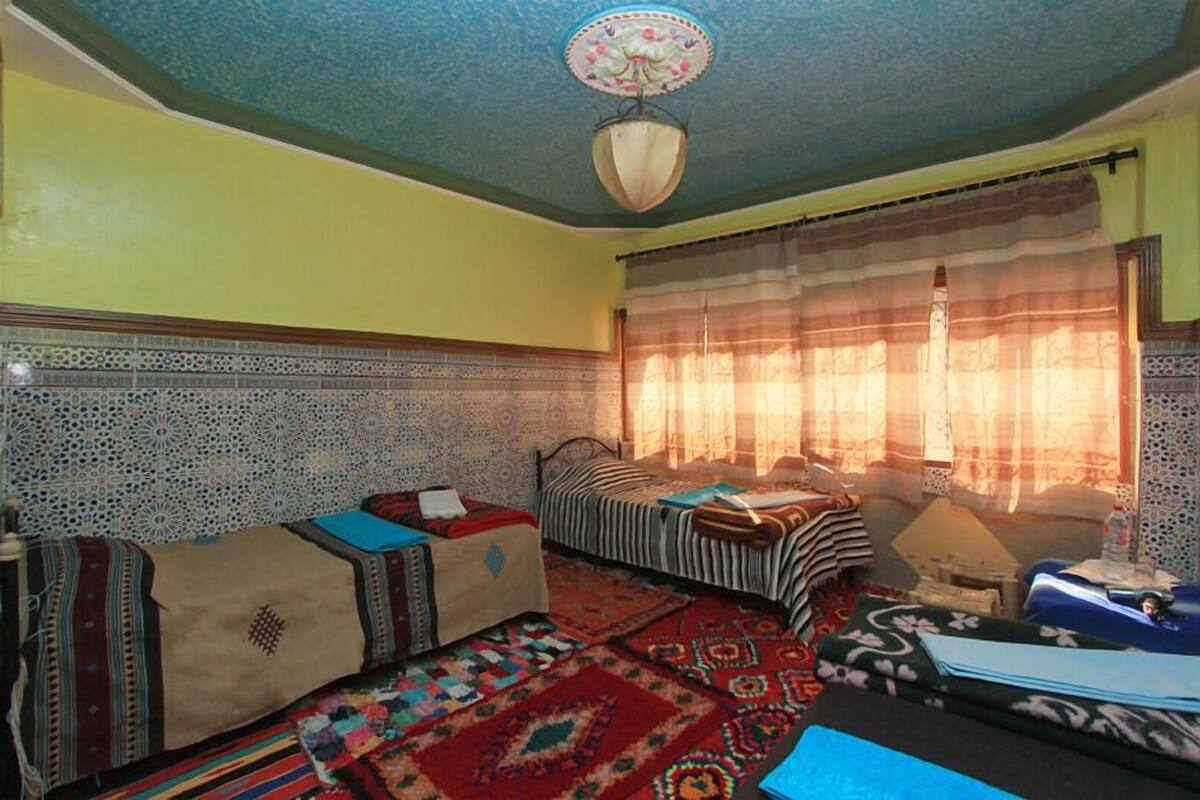 A room with a bed, a sofa, and a small table. There is a window with curtains and a ceiling fan.
