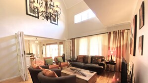 A spacious living room with a high ceiling, a chandelier, a large window with curtains, and a fireplace. - Jonquil Cottage (Franschhoek)