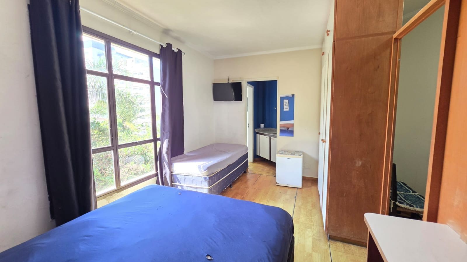 Ein Zimmer mit einem Bett, einer kleinen Küchenzeile, einem Fernseher und einem fenster mit Vorhängen.