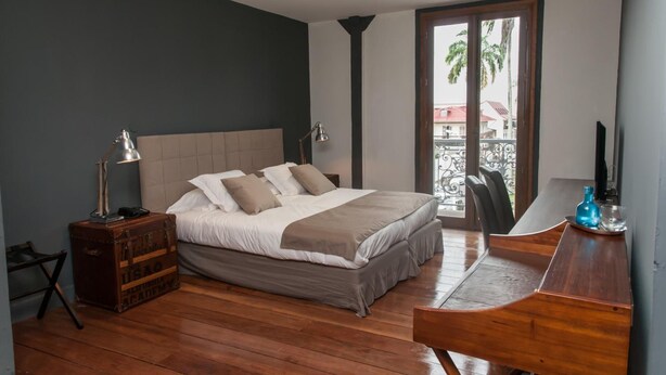 A bedroom with a bed, bedside lamps, a wooden desk, and a suitcase. - Hôtel des Palmistes (Cayenne)