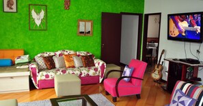 A living room with a green wall, a floral sofa, a pink armchair, a wooden guitar, and a television displaying a guitar player. - Curitiba Casa Hostel (Curitiba)