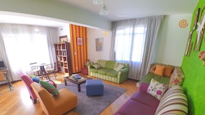 A living room with a green sofa, a purple sofa, a blue ottoman, and a red armchair. There is a bookshelf, a coffee table, and a dining table with chairs. - Curitiba Casa Hostel (Curitiba)