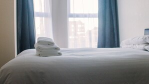 Een net opgemaakt bed met een stapel witte handdoeken, een wit kussen en een raam met blauwe gordijnen.