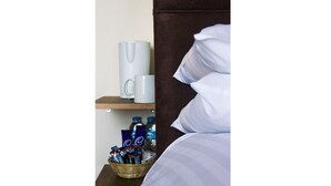 Hotelkamer met een bed, nachtkastje met een witte mok, verzorgingsset en plank met waterflesjes en een blauw voorwerp.