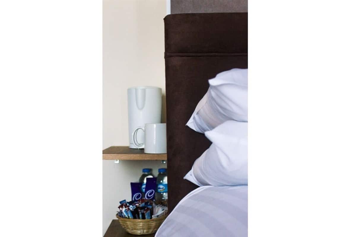 A hotel room with a bed, a bedside table with a white mug, a basket of toiletries, and a shelf with bottled water and a blue item.