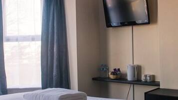 A hotel room with a bed, a flat-screen TV mounted on the wall, a window with curtains, and a small table with toiletries.