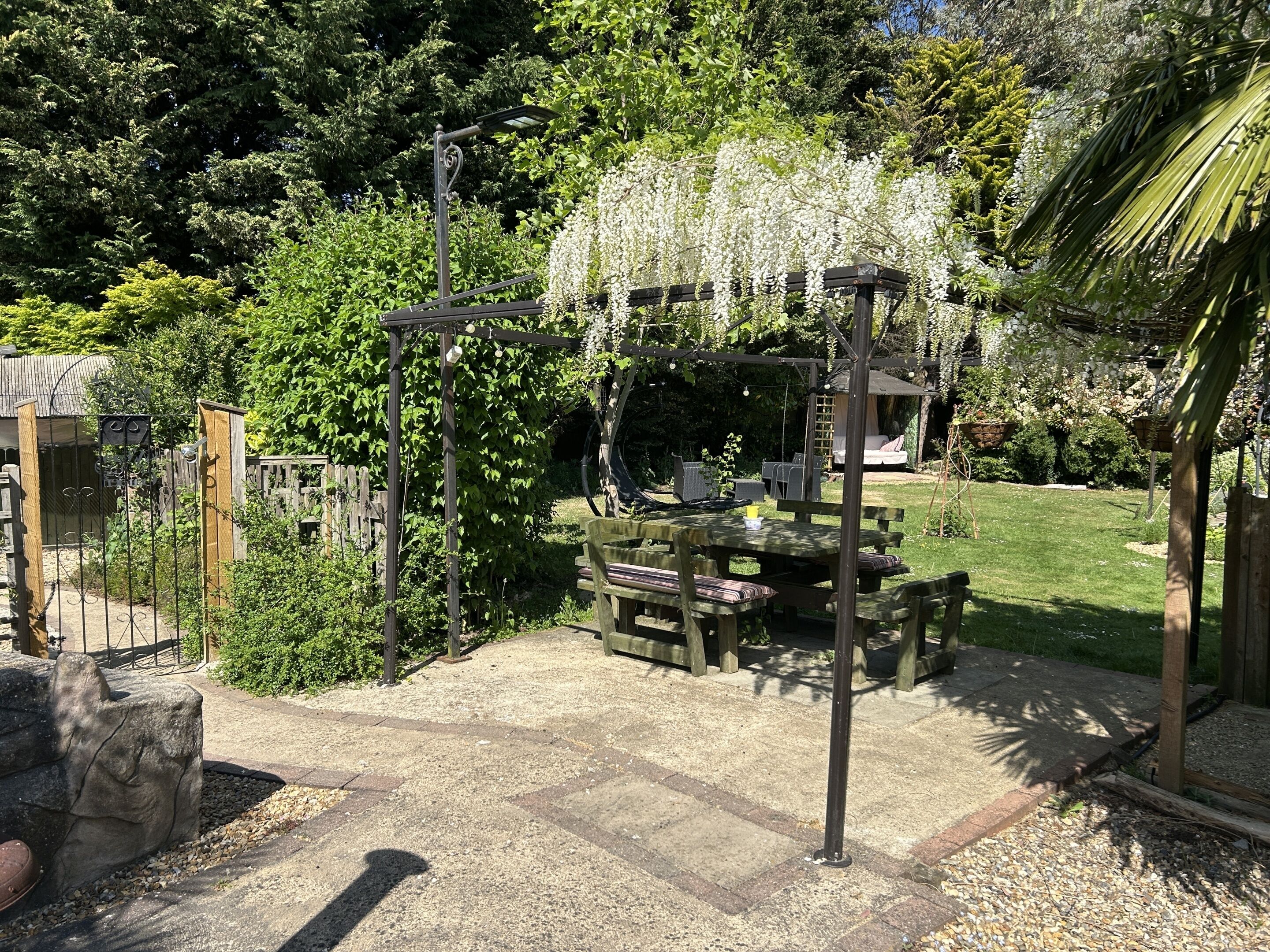 Un patio con mesa y sillas, rodeado de vegetación y una pérgola con luces colgantes.