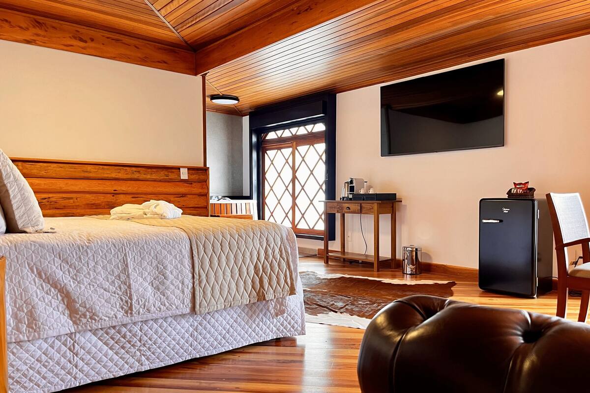 A bedroom with a bed, wooden ceiling, a desk, a chair, a small refrigerator, and a window with a view.