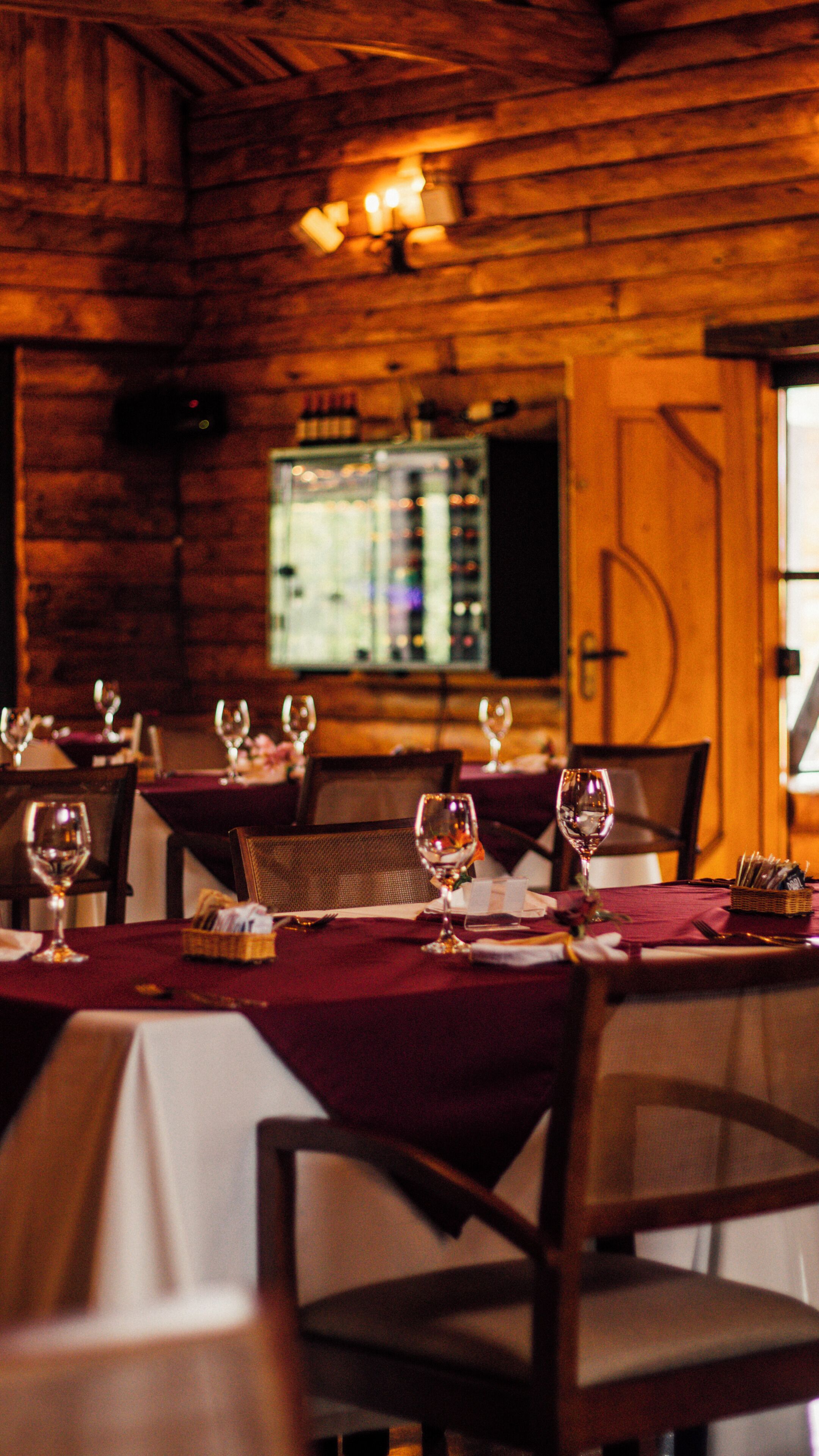 A restaurant interior with wooden walls, tables set with wine glasses, and a bar area in the background.