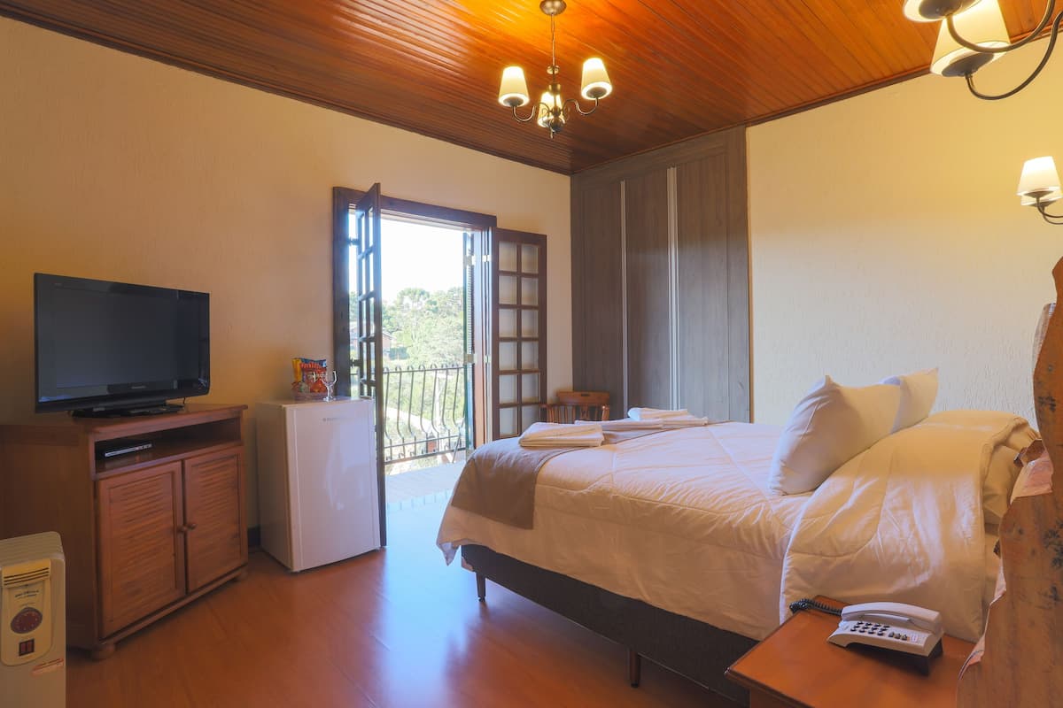 A hotel room with a bed, a television, a refrigerator, a wooden cabinet, and a balcony with a view of trees.