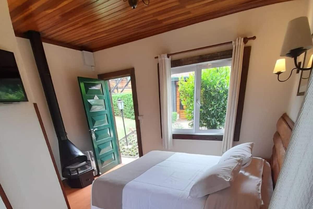 A bedroom with a bed, a bedside table with a phone and remote, a window with curtains, and a door leading outside.