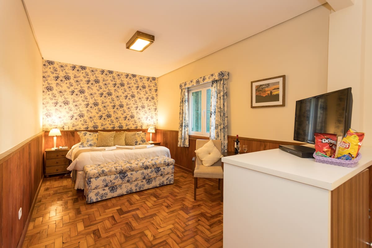 A bedroom with a wooden herringbone floor, a bed with floral bedding, a nightstand, a chair, a television, and a basket of snacks.
