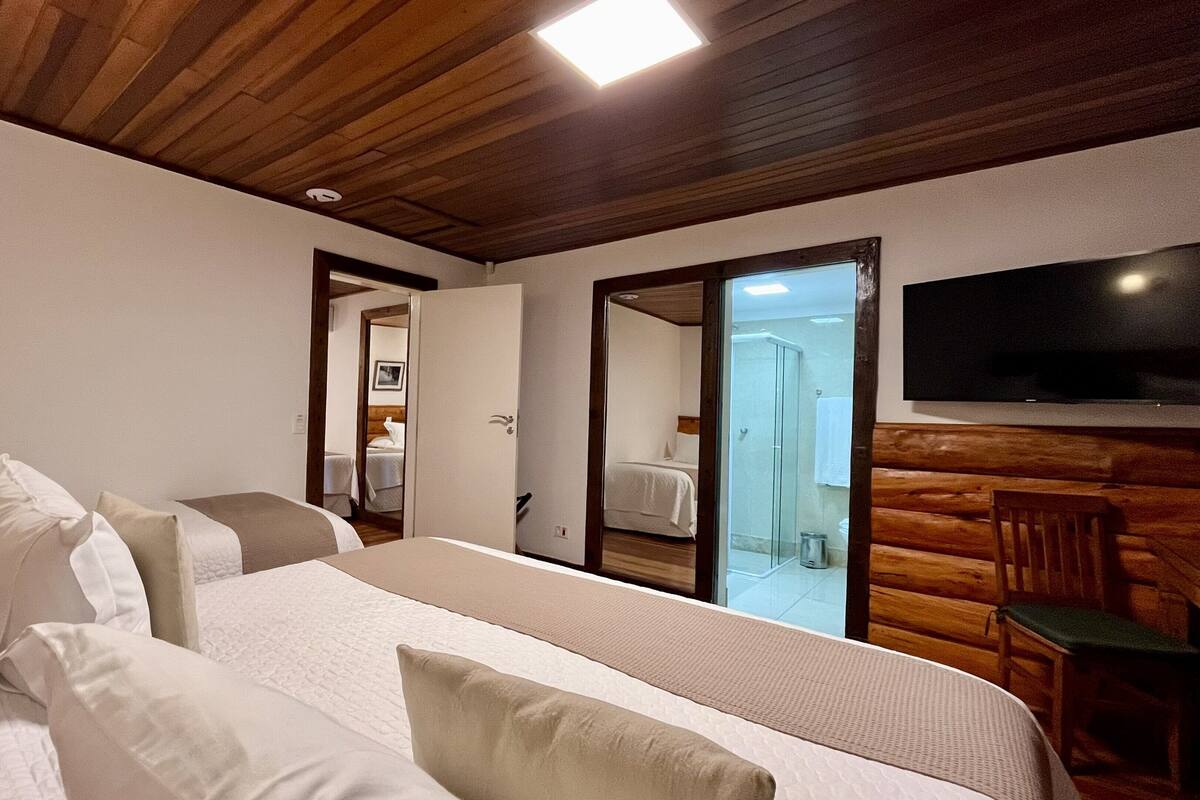 A hotel room with a wooden ceiling, a large bed with white bedding, a wooden bench, and a TV mounted on the wall.