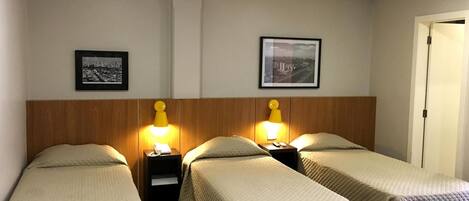 A hotel room with two single beds, each with a wooden headboard and a bedside table with a lamp. There are two framed pictures on the wall above the beds.