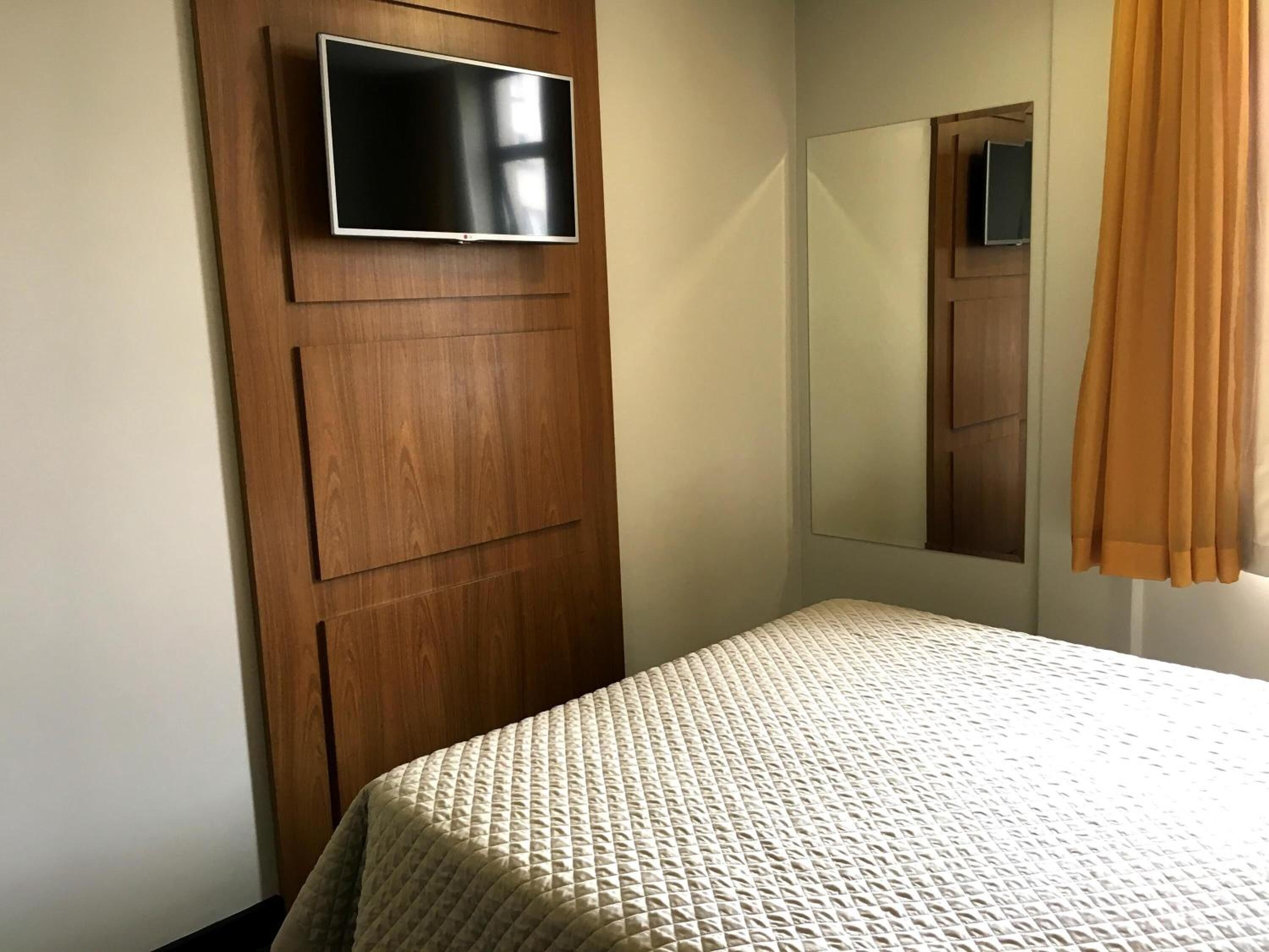 A bedroom with a bed, a TV mounted on a wooden panel, a mirror, and a window with curtains.