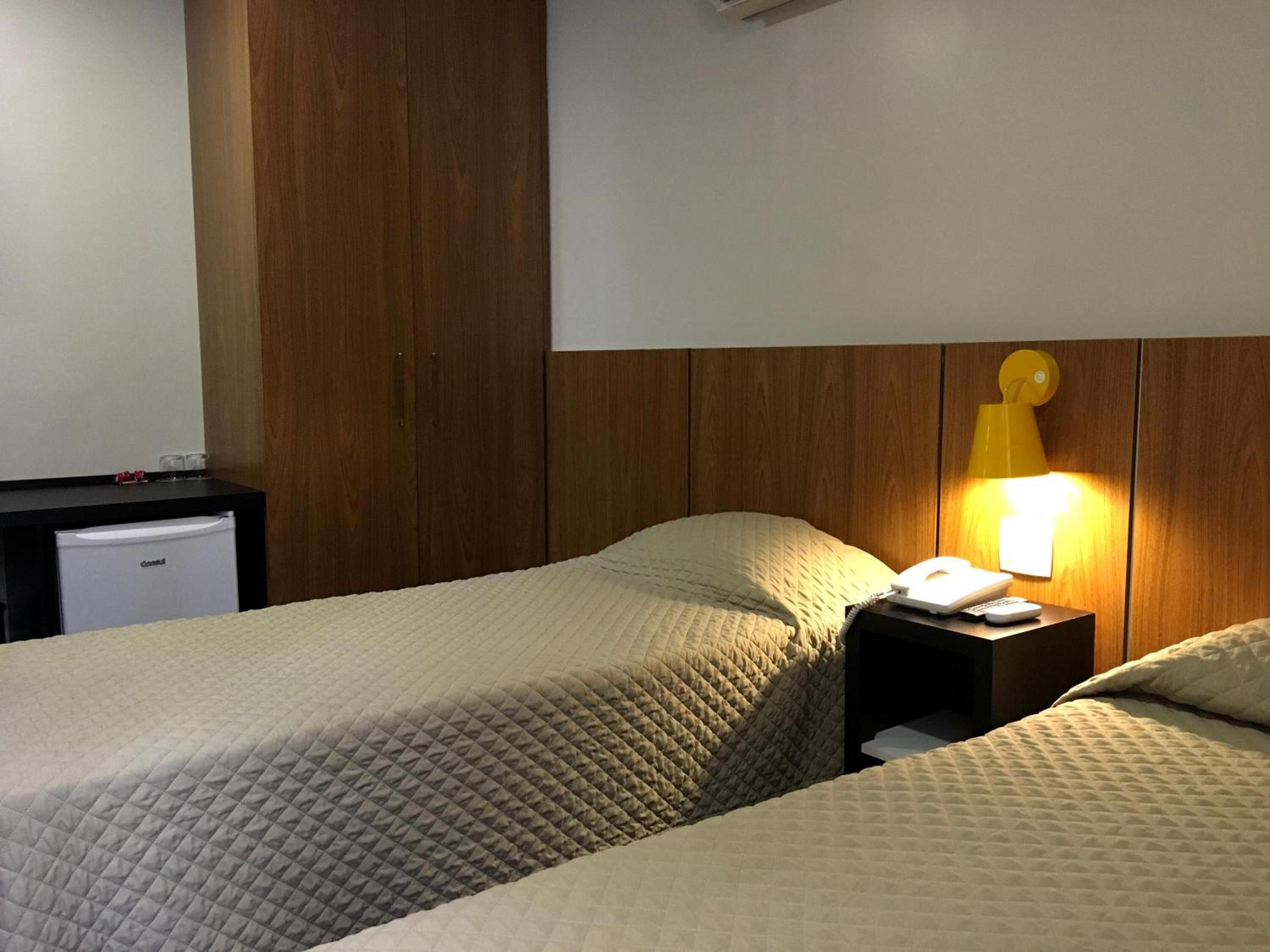 A hotel room with two beds, a bedside table with a lamp, a small desk with a phone, and a minibar.