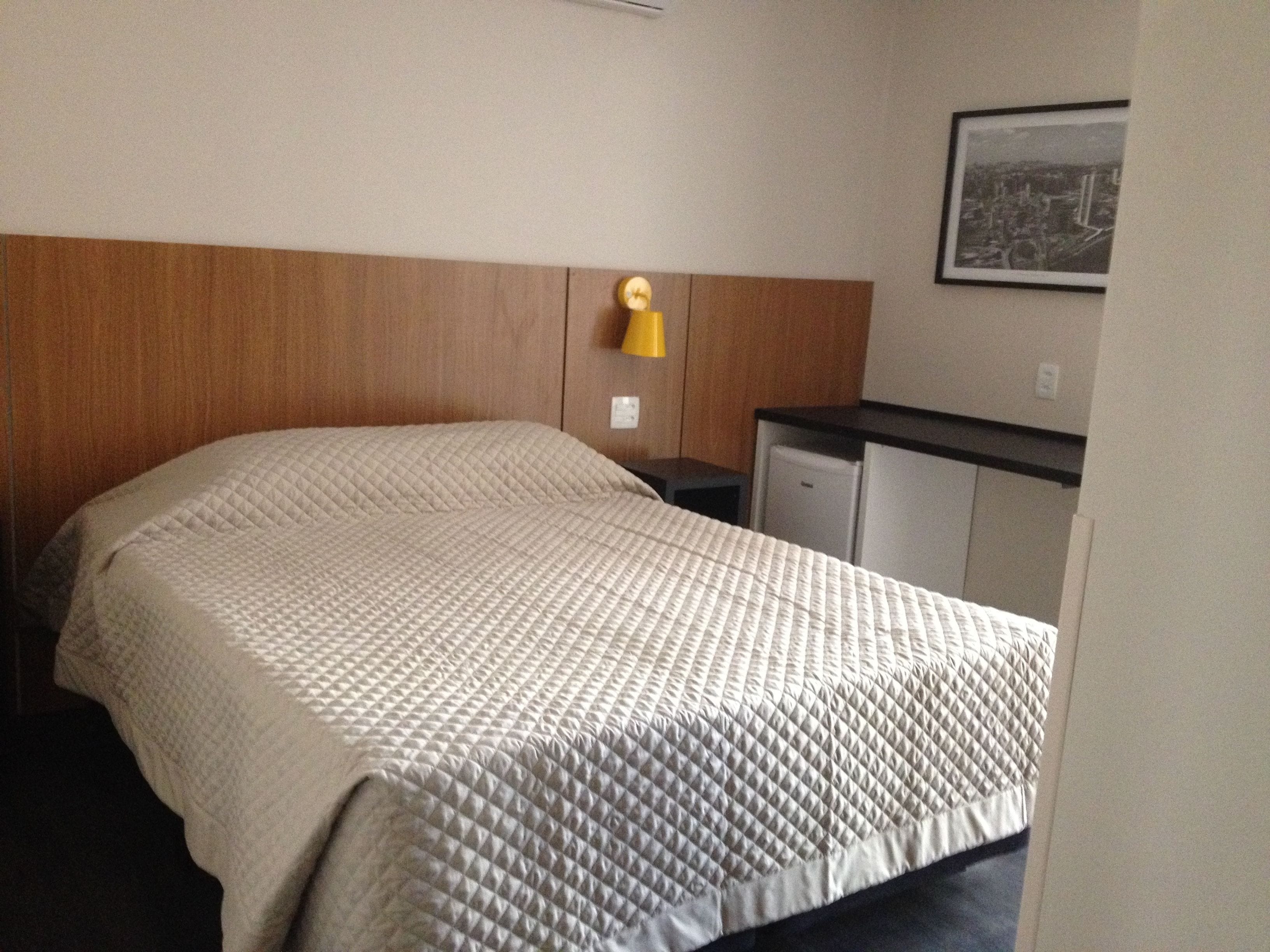 A hotel room with a bed, a small desk, a refrigerator, and a wall-mounted lamp.