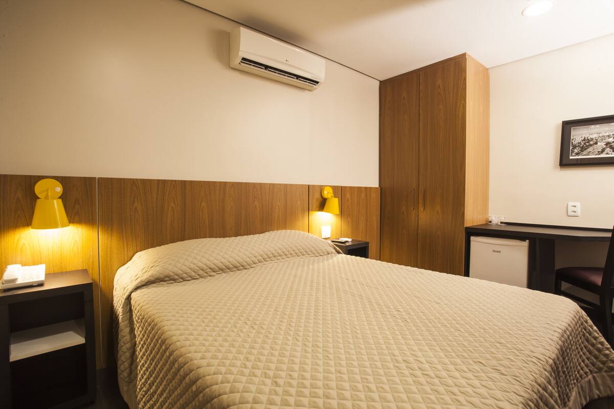 Ett hotellrum med en säng, sänglampor, ett skrivbord och ett litet kylskåp.