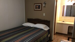 Una cama individual con una colcha a rayas y dos almohadas, un pequeño cuadro en la pared, y un baño con lavabo y espejo.
