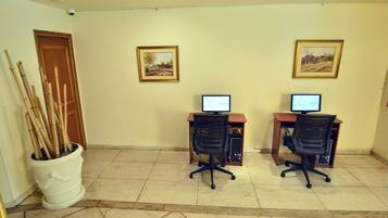 A room with two desks, each with a computer and a chair. There are two framed pictures on the wall. A white pot with bamboo sticks is on the floor to the left.