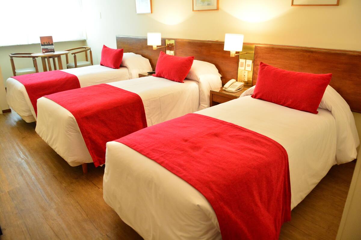 A hotel room with three beds, each with white bedding and red pillows. There are bedside tables with lamps and a small table with a sign.