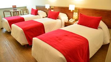 A hotel room with three beds, each with white bedding and red pillows. There are bedside tables with lamps and a small table with a sign.