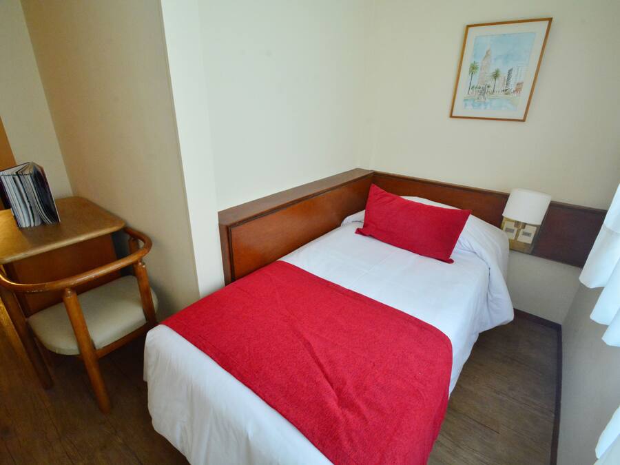 Habitación de hotel con cama de madera, almohada roja, colcha blanca, una silla de madera y una mesita.