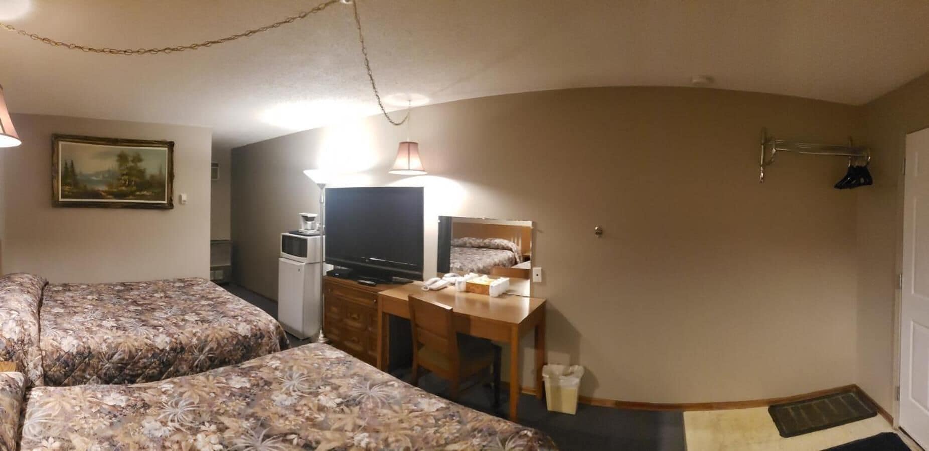 A hotel room with two beds, a desk with a television, a refrigerator, and a small table with a lamp.