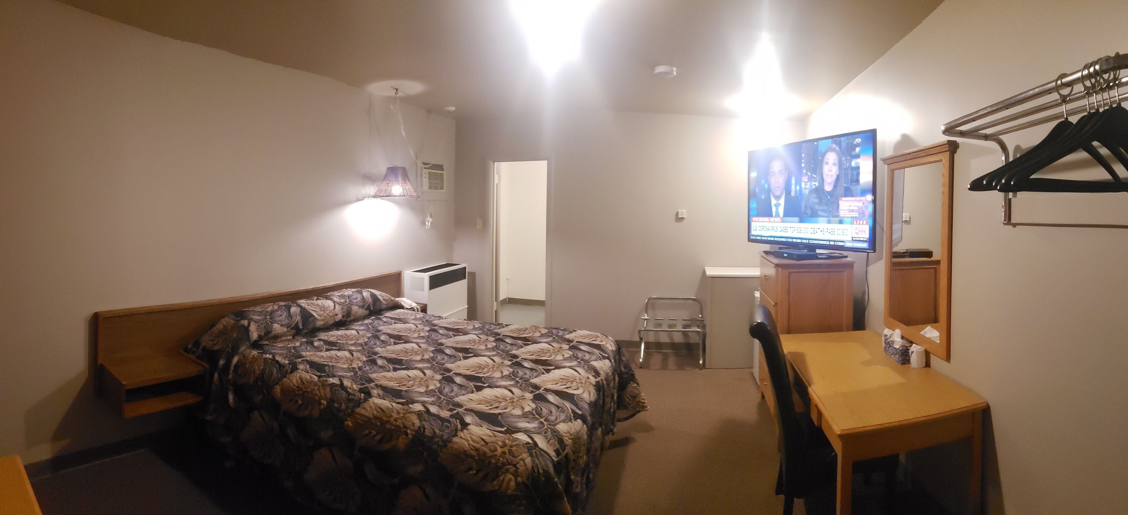 Un dormitorio con cama, escritorio, silla, televisor, espejo y perchero.