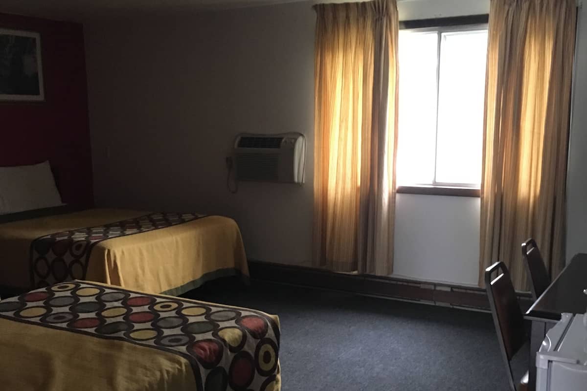 A hotel room with two beds, a desk, a chair, an air conditioning unit, and a window with curtains.