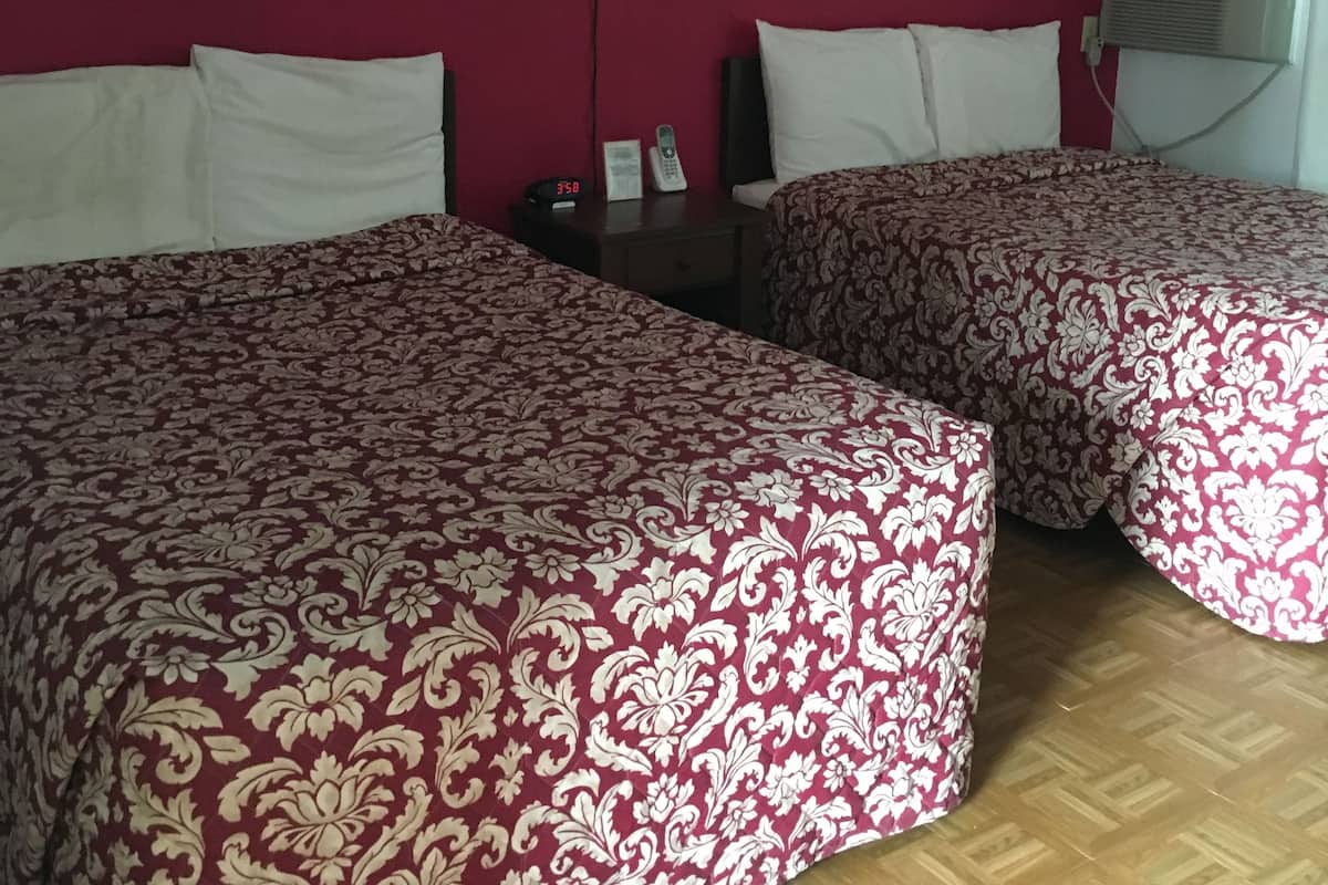 A hotel room with two beds, each with a patterned bedspread, a nightstand with a lamp, a wall-mounted air conditioner, and a red wall.