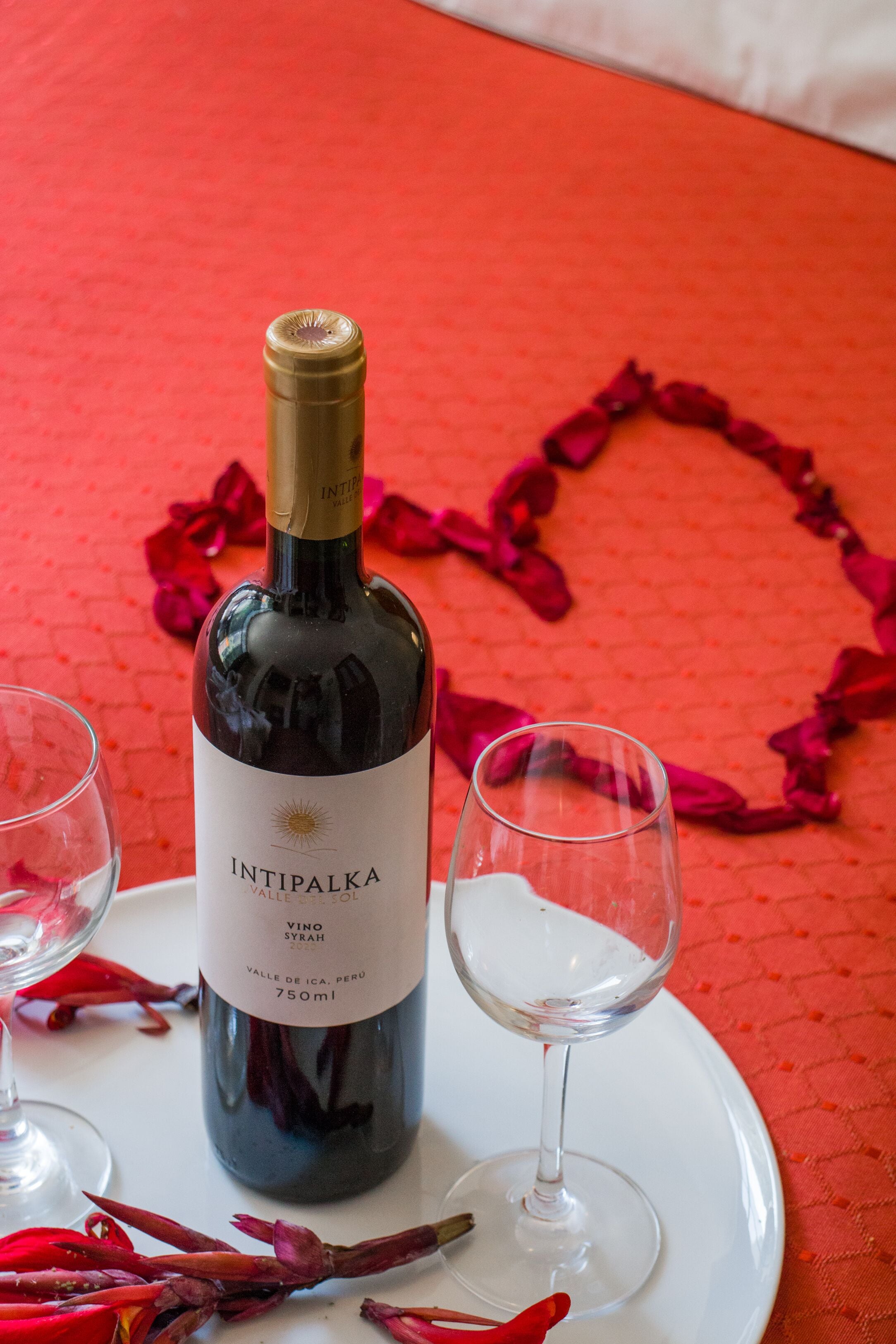 A bottle of wine, two glasses, and red rose petals arranged on a table.